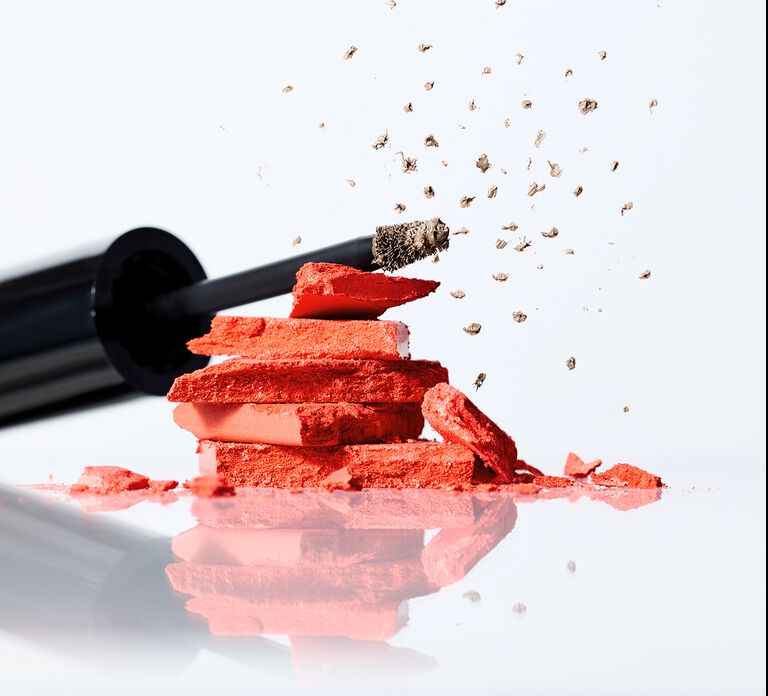 Crushed coral blush stacked in pieces with a brow‑tint applicator flicking tinted particles