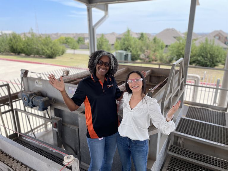 Two people standing near industrial equipment outdoors under a covered structure
