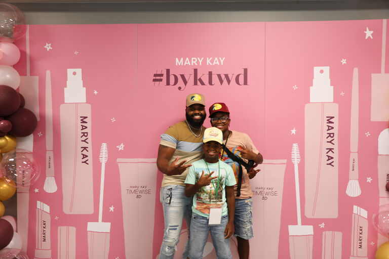 Three people posing in front of a pink Mary Kay backdrop with branded graphics and balloons